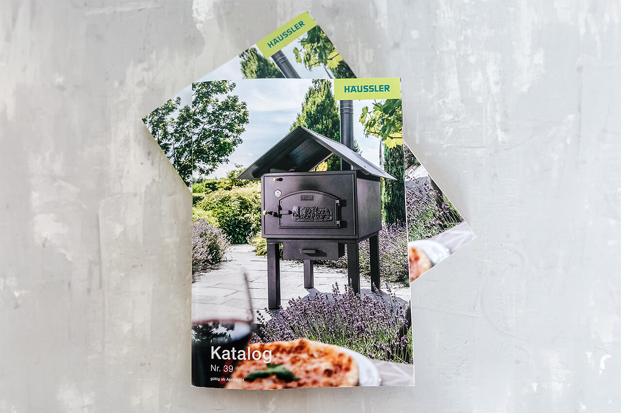 Häussler Gesamtkatalog Abbildung des Häussler Produktkatalogs: der Katalog zeigt eine ansprechende Terrasse mit Lavendelbüschen und einem Häussler Holzbackofen. Im Vordergrund ist eine frisch gebackene Pizza zu sehen.