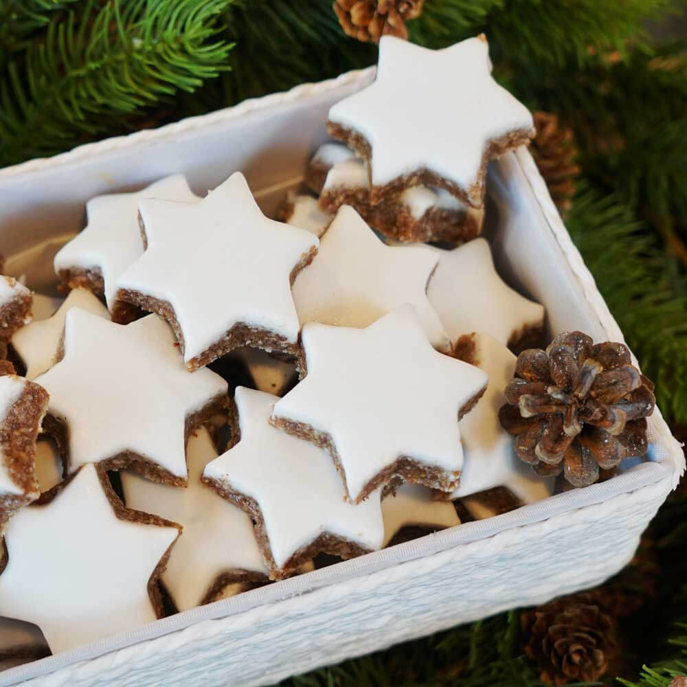 Zimtsterne mit weißer Glasur in einer weißen Box. Um die Box herum befinden sich Tannenzweige.