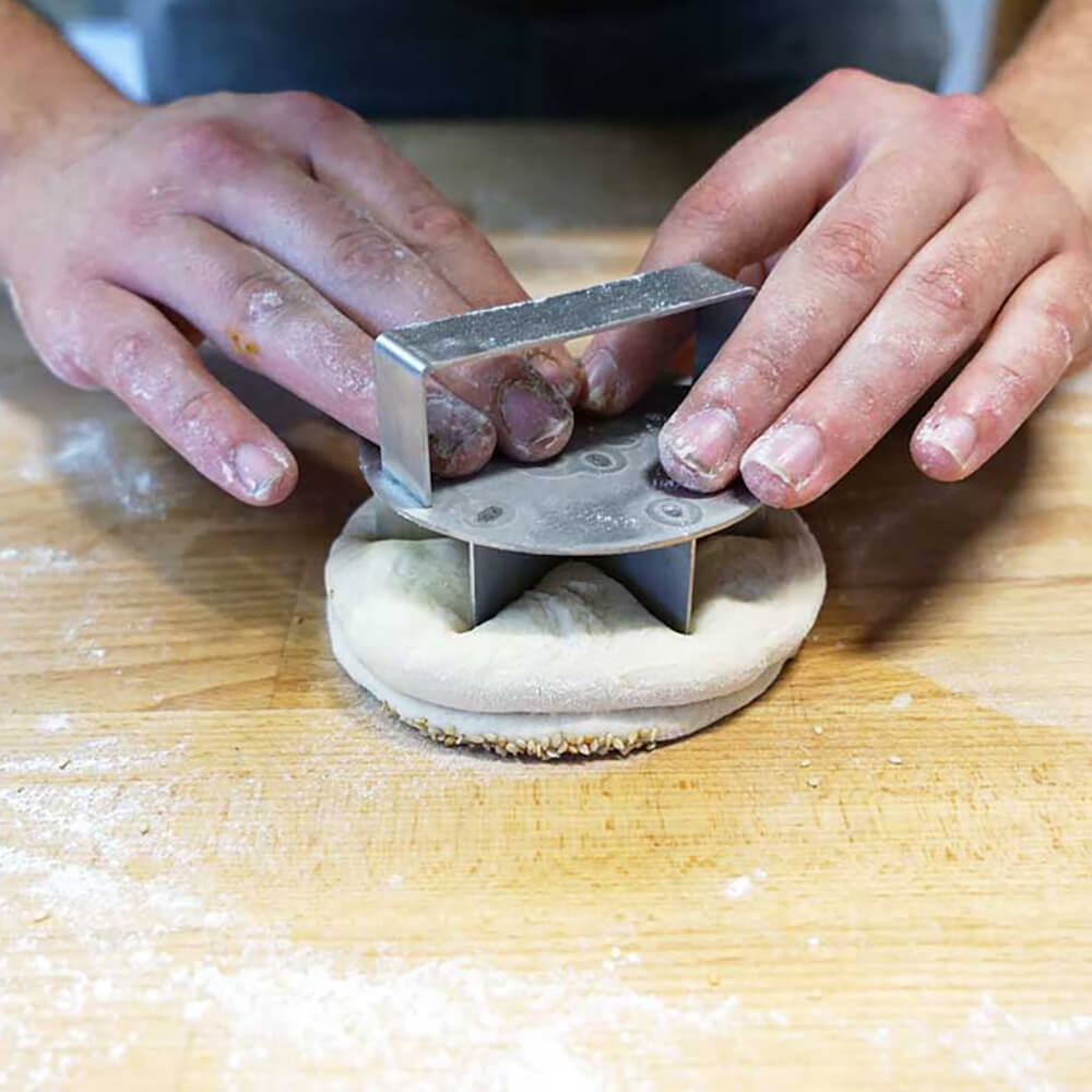 Ein Teigling liegt auf einem hellbraunen Arbeitstisch und es wird gerade mit dem Sternbrotdrücker Sternformartige Schnitte in den Teigling gedrückt