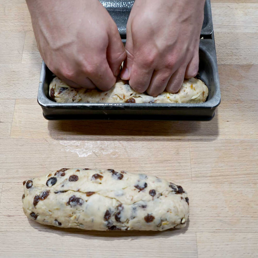 Ein Christstollen Teigling mit Cranberrys liegt auf einem braunen Tisch und daneben wird ein Christstollen Teigling mit Cranberrys in eine Christstollenform gelegt