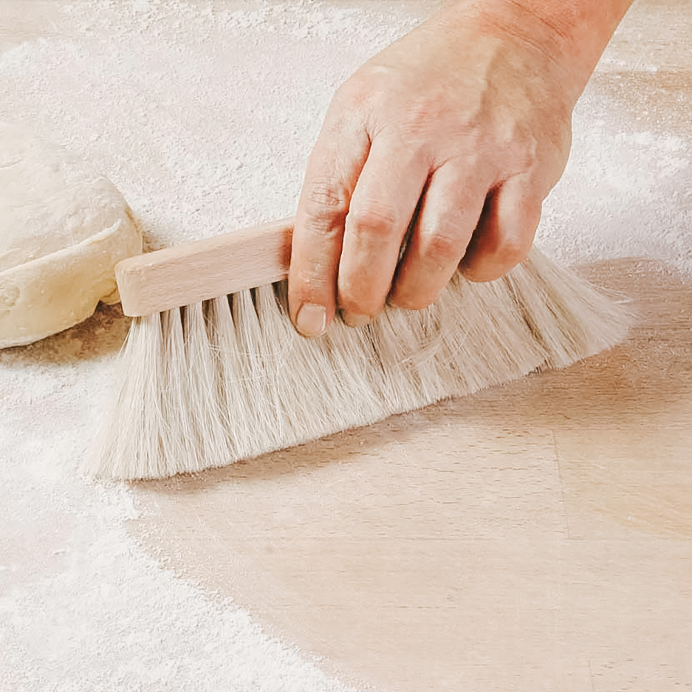 Mehlbesen 2-reihig Auf dem Bild befindet sich links ein Teigling und in der Mitte jemand der mit dem Mehlbesen Mehl auf einer hellbraunen Arbeitsplatte zusammenkehrt