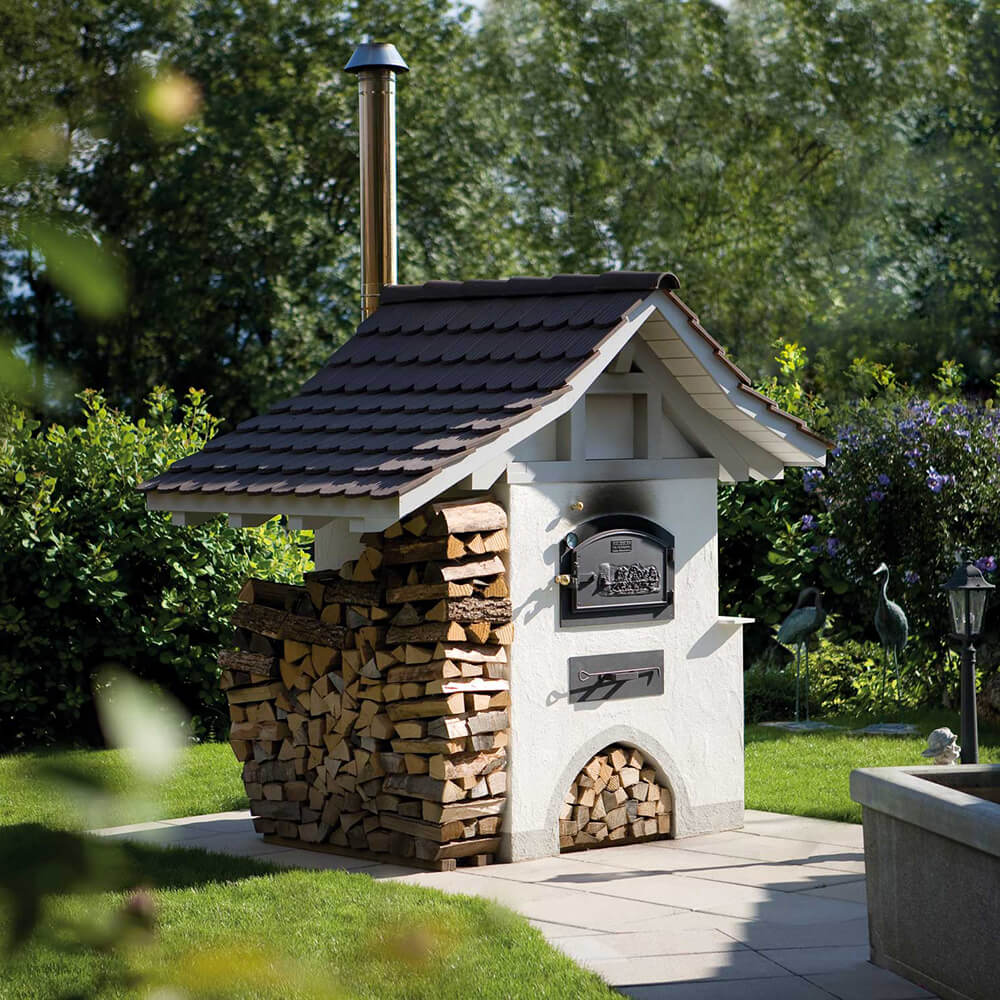 Bausatz HABO - Türmodell B Aufgebauter HABO Bausatz zu einem schönen Backhäuschen mit einigen Bäumen im Hintergrund und gestapeltem Holz neben dem Backhäuschen.