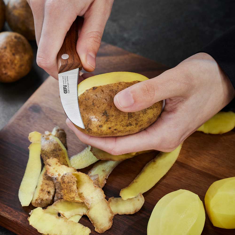 Eine Person schält mit dem Schälmesser Kartoffeln. Auf dem darunter liegenden Holzbrett liegen Kartoffelschalen.