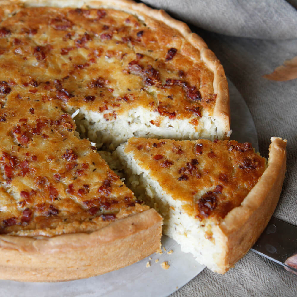Rezept für Zwiebelkuchen Zwiebelkuchen auf einem grauen Kuchenheber bei dem gerade ein Stück herausgeschnitten wird