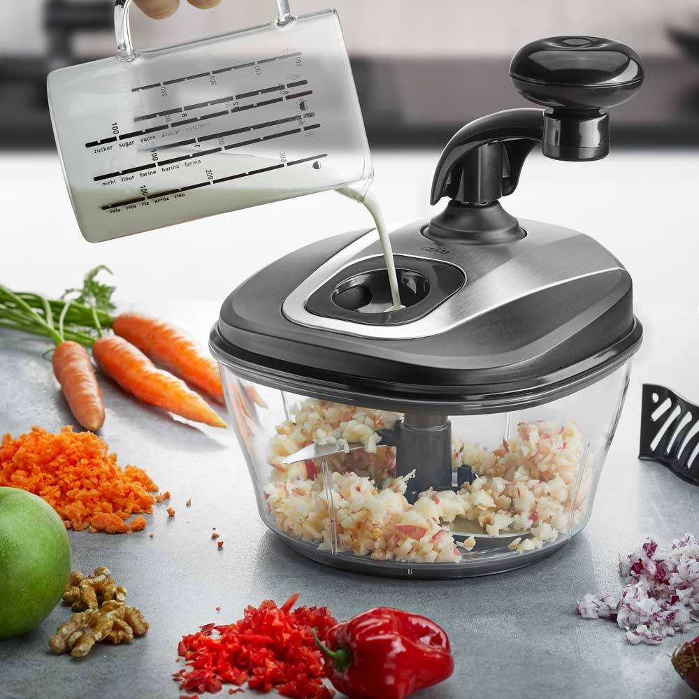 Multizerkleinerer SPINO Multizerkleinerer mit gehaktem Gemüse auf einer Küchenarbeitsplatte. Rings um liegt verschiedenes Gemüse, jemand gießt Flüssigkeit ein.