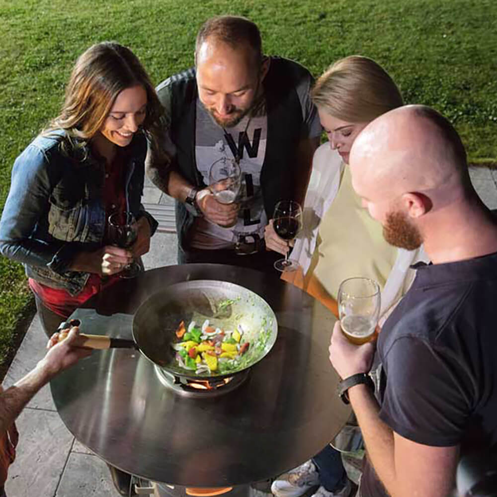 Wok Stahl Vier Menschen stehen in einem Garten um ein Ambientelicht, auf welchem der Grillrost aufgelegt ist. Auf dem Rost befindet sich der Wok mit Grillgemüse.