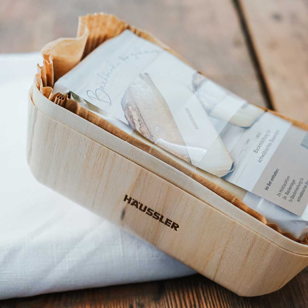 Holzbackform-Set von Häussler – Nahaufnahme Nahaufnahme eines Holzbackform-Sets von Häussler, mit Backmischung für Brot, in einer rechteckigen Holzform auf einem weißen Stoff liegend