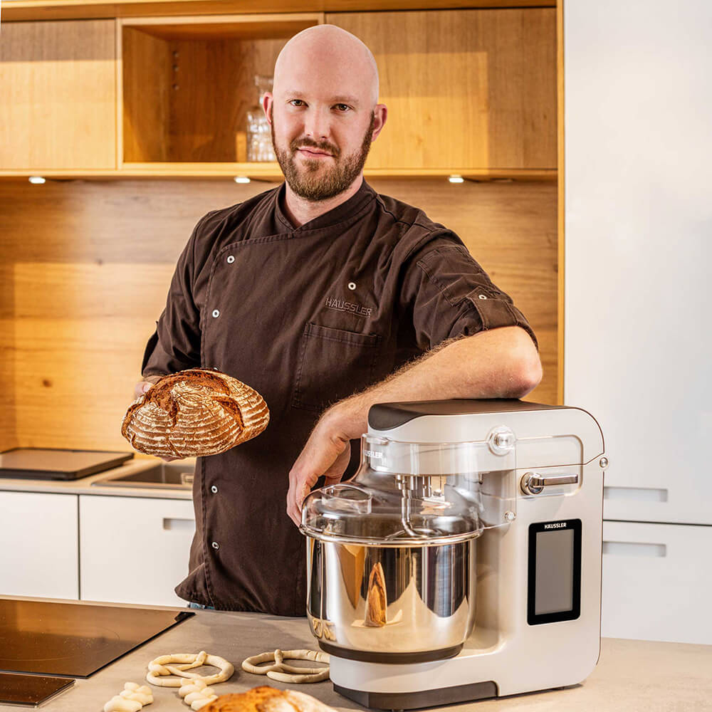 Ein Bäcker mit einem Brot in der Hand legt in der Küche seinen Arm auf der NOVA Teigknetmaschine ab.