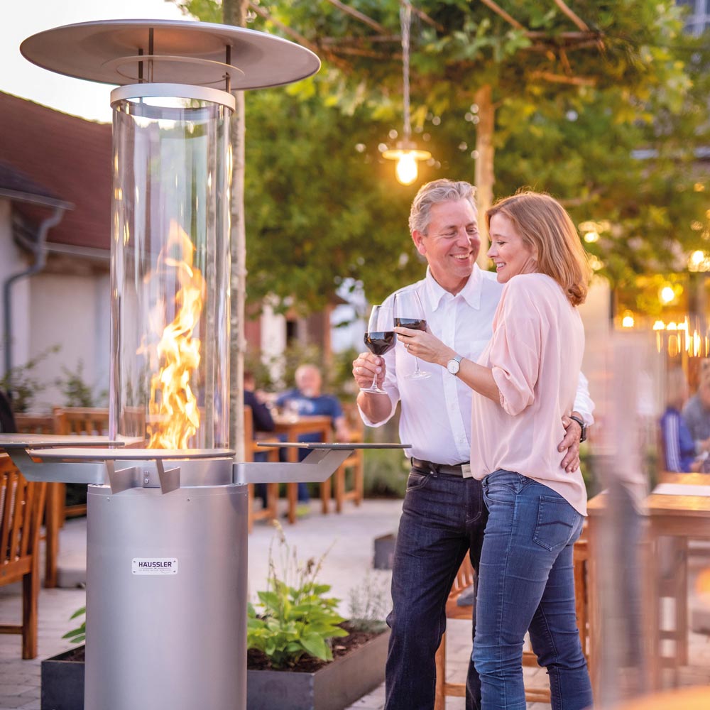 Gastro-Paket Terrassenstahler Terrassenstahler mit angezündetem Feuer. Danben steht ein Paar mit einem Glas Wein unter einem Baum und schaut den Terrassenstrahler an.