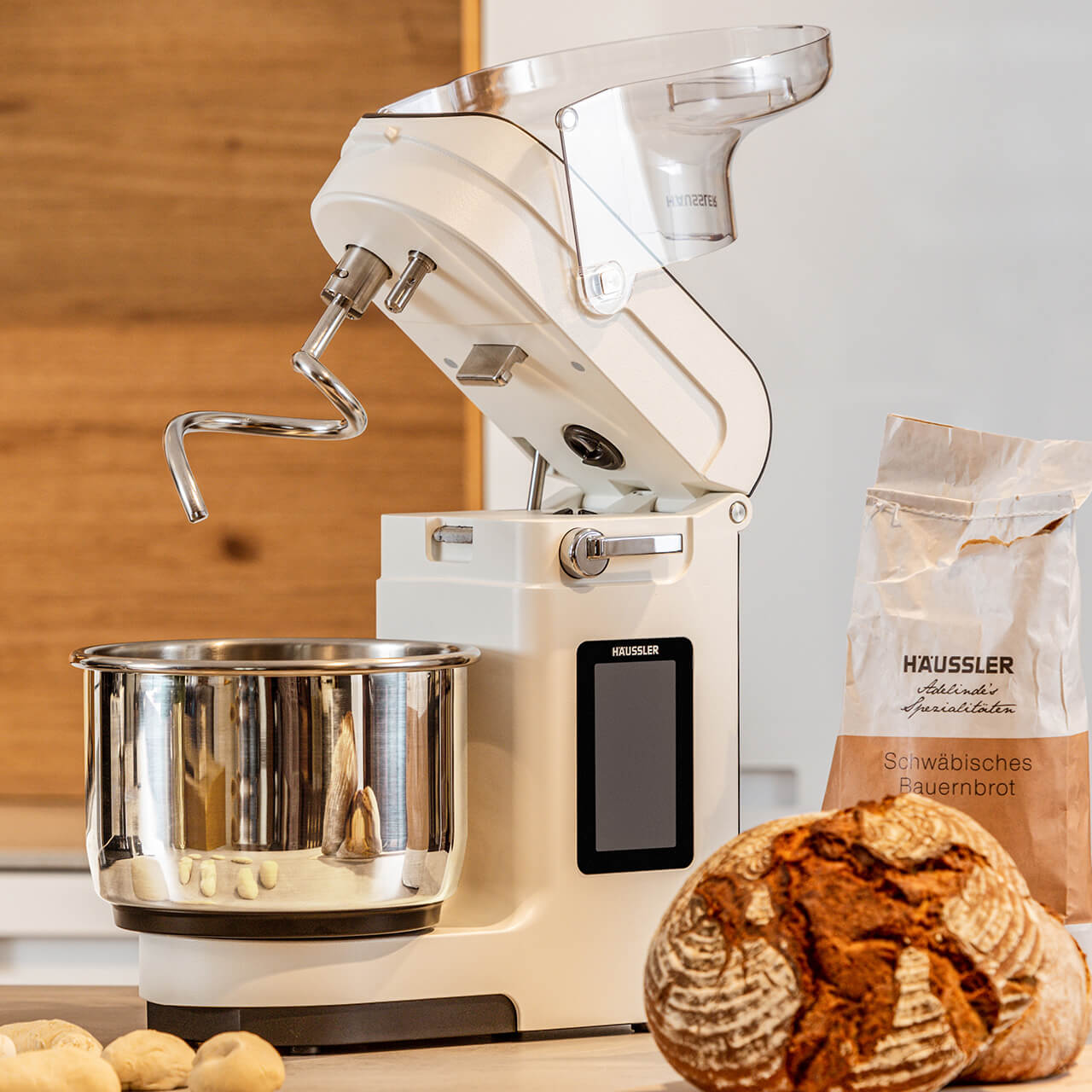 Teigknetmaschine_Nova_mit_Brot Abbildung einer NOVA Teigknetmaschine in weiß vor einer Holzwand. Im Vordergrund steht einen Mehltüte sowie ein frisch gebackenes Brot.