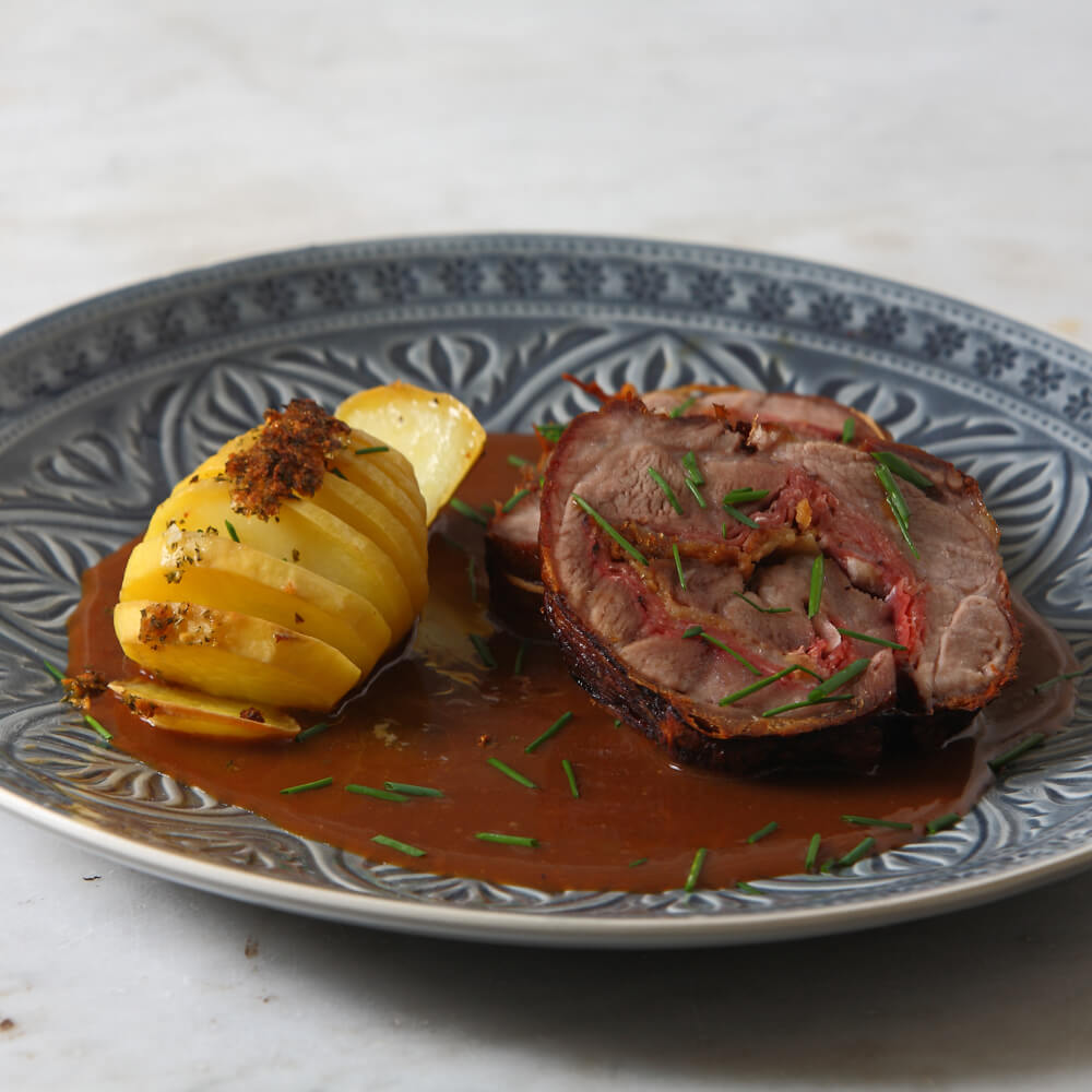 Rezept für Gefülltes Schweinekotlett mit Rosmarinkartoffeln und gefüllten Tomaten gefülltes Schweinekotlett mit einer gefächerten Rosmarinkartoffel, einer braunen Soße und Schnittlauch auf einem Blau gemusterten Teller
