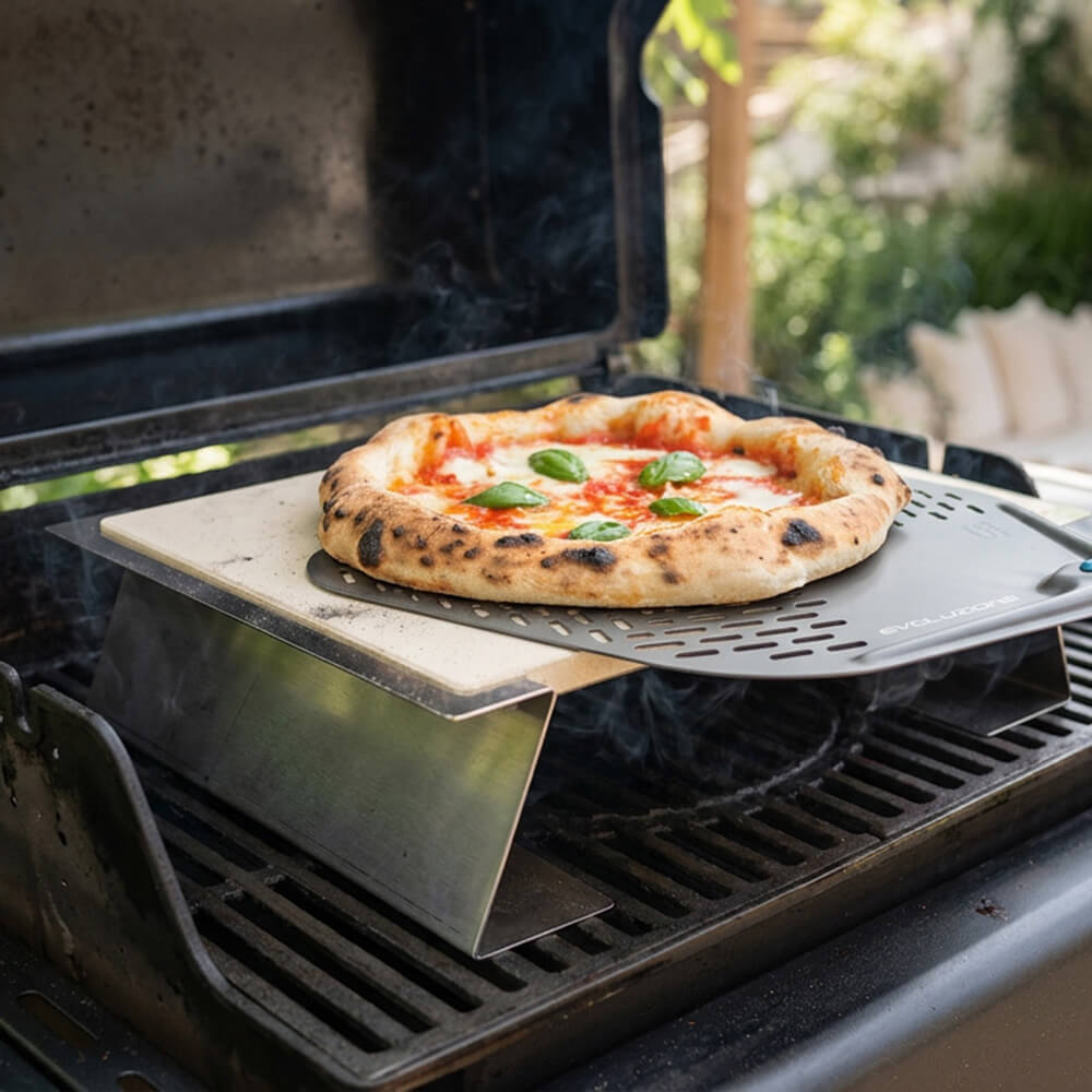 Pizzastein mit Halterung für den gasgrill Pizzastein auf Halterung auf Grillrost. Darauf ist eine gebackene Pizza zu sehen, die von einem Backschieber heruntergenommen wird.. Im Hintergrund eine Terrassenumgebung