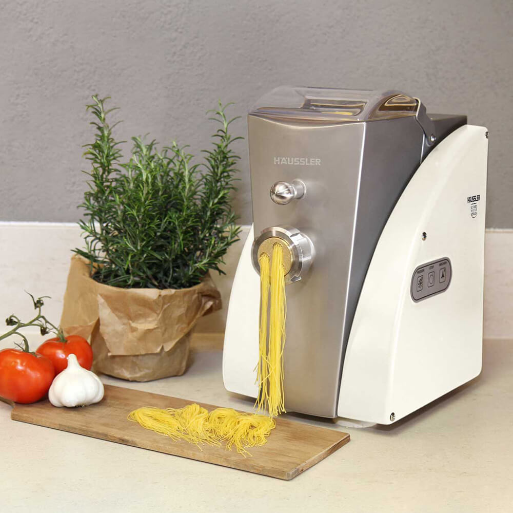 Nudelmaschine LUNA Cremeweiß Nudelmaschine Luna in der Farbe cremeweiß beim herstellen von frischen Nudeln. Im Hintergrund sind noch ein paar Tomaten, Knoblauch und ein Strauch Rosmarin zu sehen