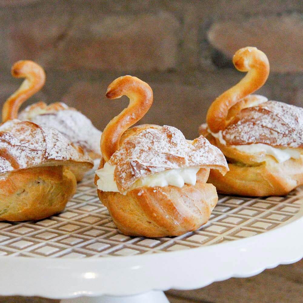 Drei Windbeutel in Schwan Form, mit Sahne gefüllt, stehen auf einer karierten Kuchenplatte.