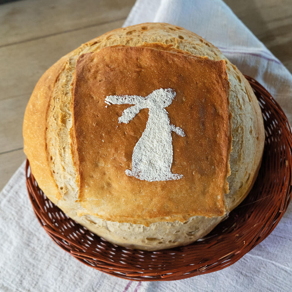 Ein Brotlaib liegt in einem geflochtenen Korb, der auf einem Tuch steht. Auf dem Brot ist ein Bild aus Mehl zu sehen in Form eines stehenden Hasen.