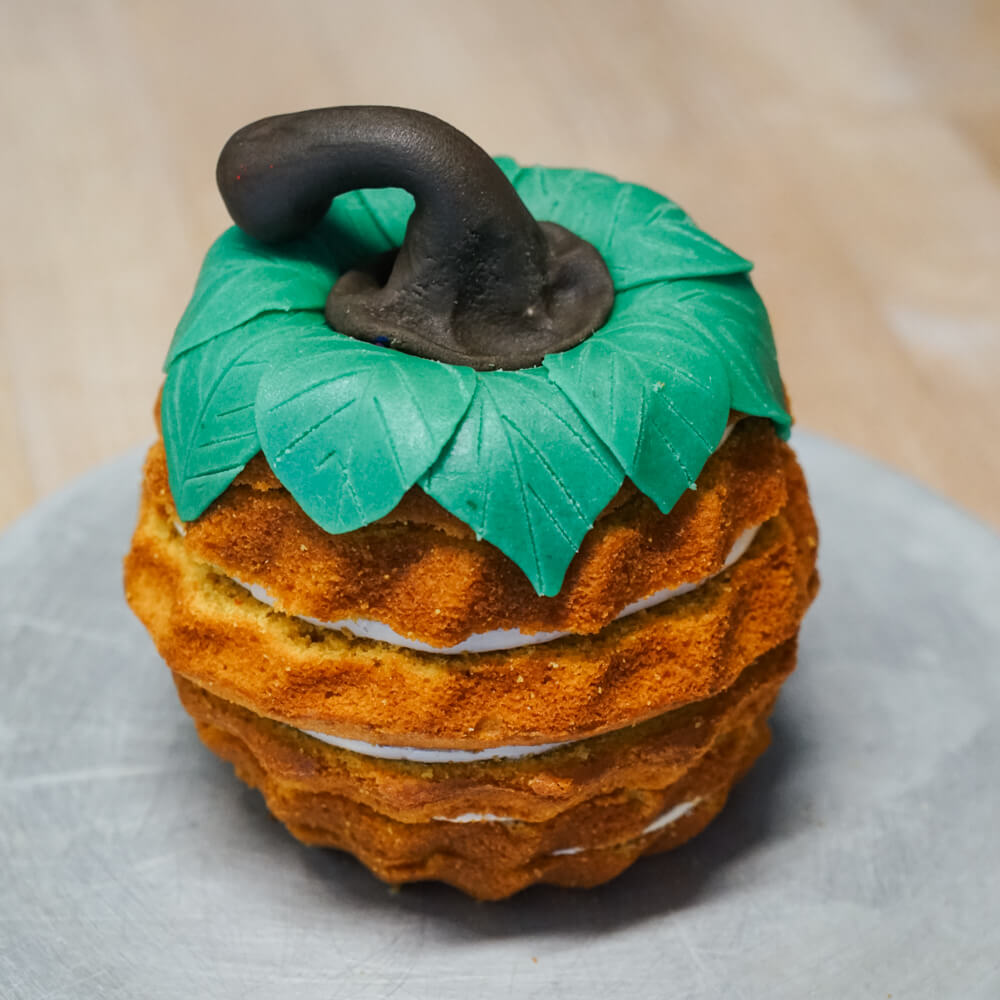 Rezept für Kürbiskuchen Kuchen in Kürbisform mit einer Creme geschichtet. Oben Drauf sind grüne Dekoblätter und ein Stiel sodass der Kuchen wie ein Kürbis aussieht