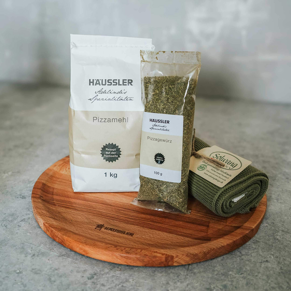 Geschenk-Set zum Verschenken und direkt los backen! Geschenk-Set mit Pizza-Mehlmischung, Pizzagewürz, Geschirrtuch und Pizza-Servierholzbrett vor einem Steinhintergrund