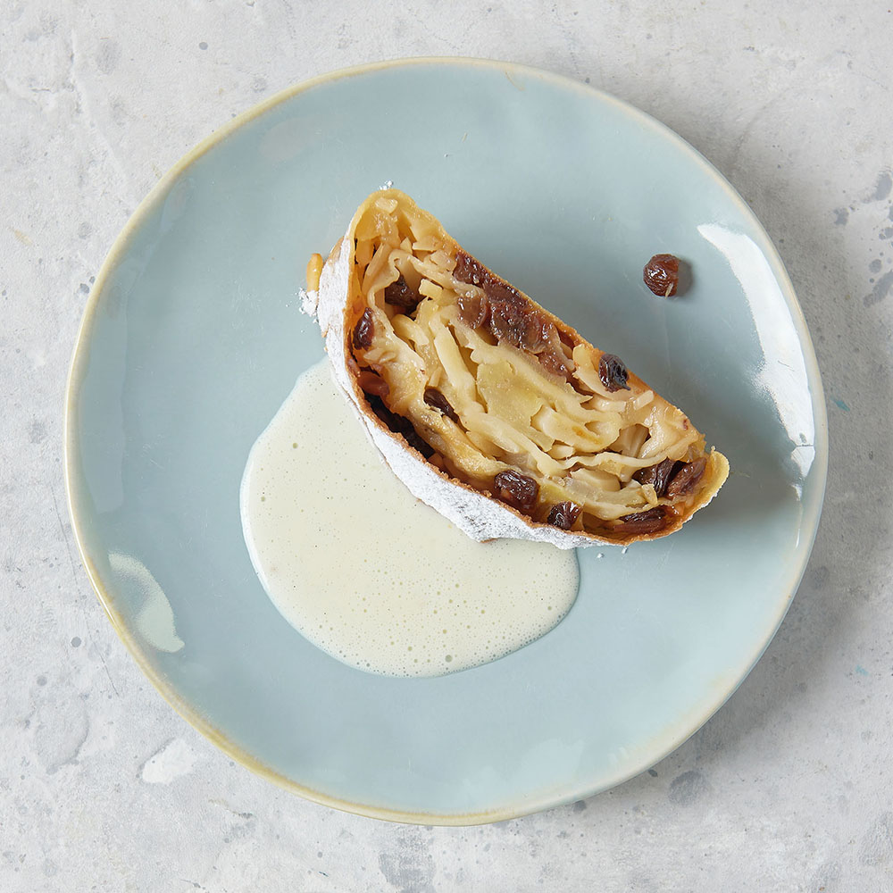 Rezept für Apfel- und Topfenstrudel ein Stück Apfelstudel mit einer Füllung aus Äpfeln und Rosinen, mit ein wenig Vanillesoße auf einem hellblauen Teller serviert