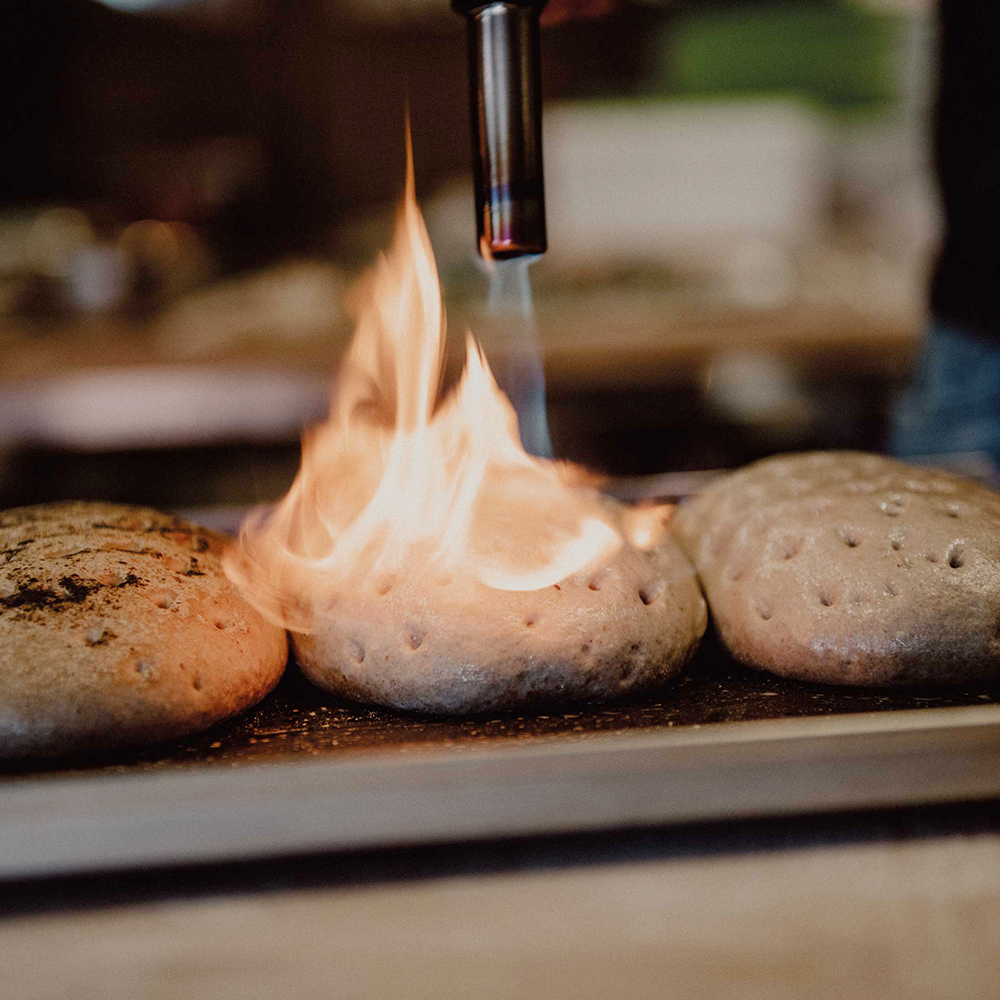 3 Pilze auf einem Edelstahlblech mit Dauerbackfolie liegend, die mit einem Flambierbrenner mir offener Flamme flambiert werden