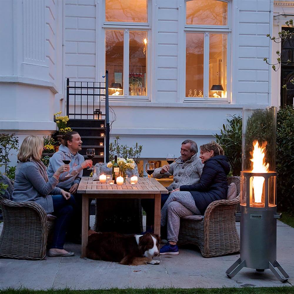 Ambientelicht XL Ein Ambientelicht mit angezündetem Feuer, daneben sitzen vier Personen und trinken ein Glas Wein.