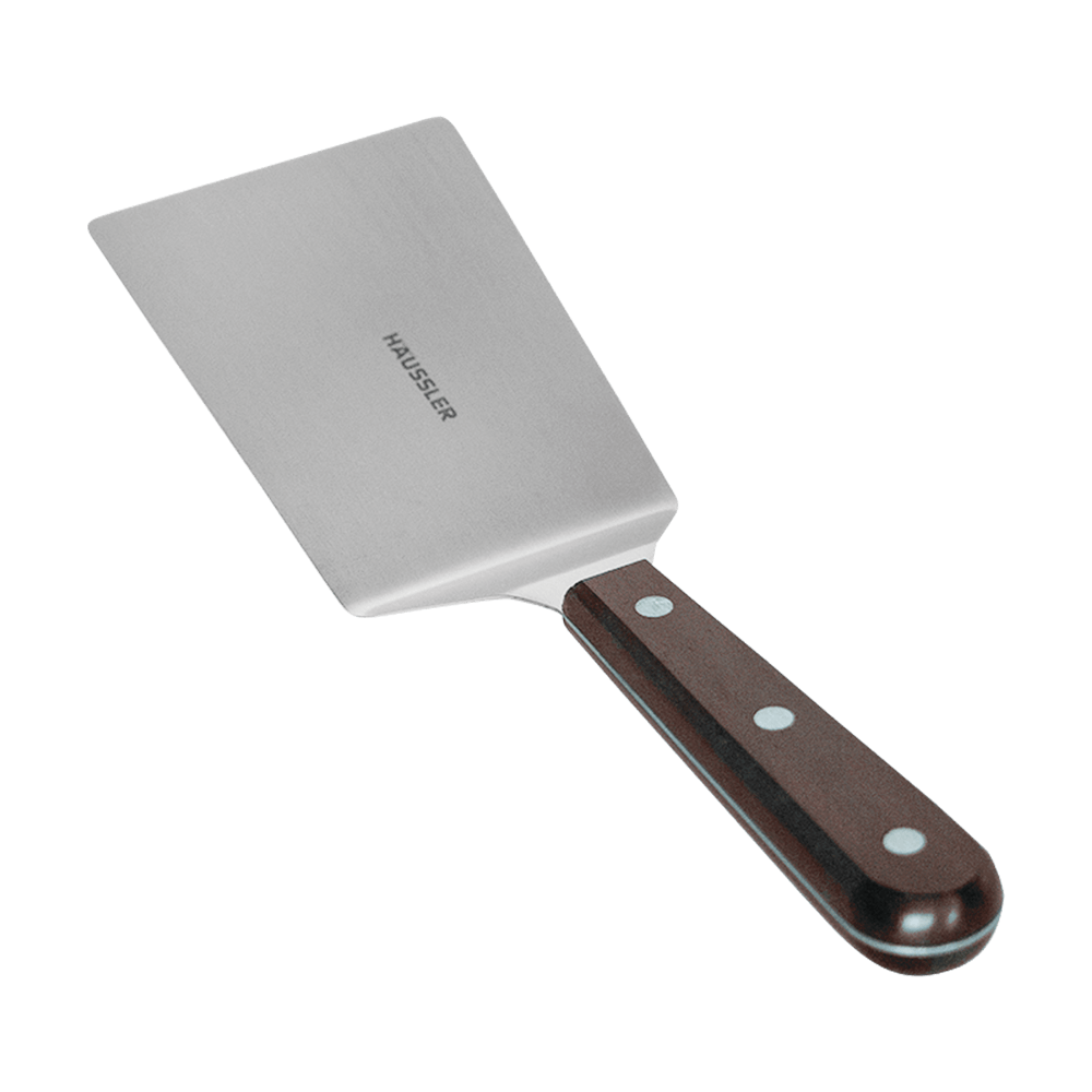 Grillpalette gekröpft Breite, eckige Grillpalette aus Edelstahl mit dunkelbraunen Griff und einem Häussler Logo auf dem Edelstahl. Ohne Hintergrund abgebildet.