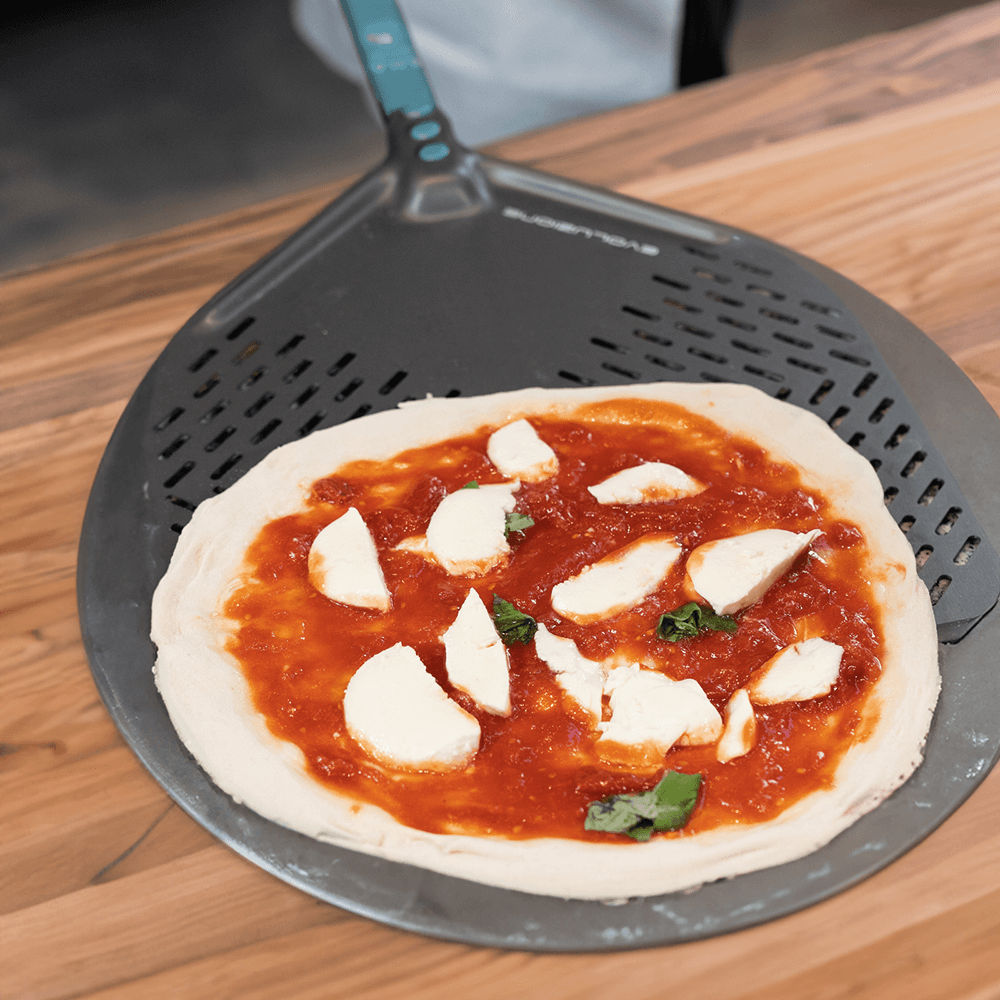 Pizzaschieber perforiert Pizzaschieber mit belegter Pizza. Die Pizza ist noch nicht fertig gebacken, belegt mit Tomatensoße, Mozzarellastücken und Basilikum.