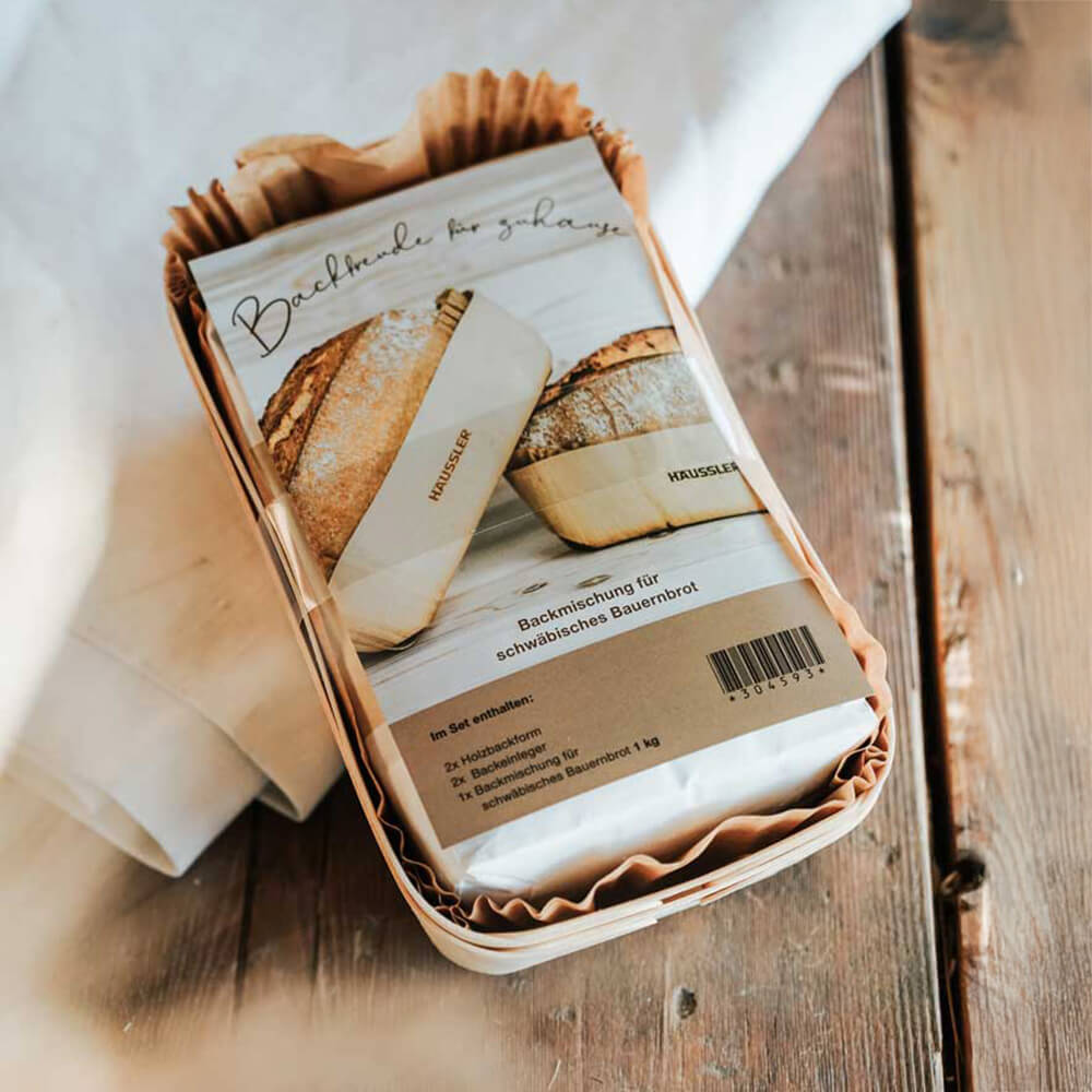 Holzbackform-Set mit Bauernbrot Backmischung Holzbackform-Set von Häussler mit Backmischung für schwäbisches Bauernbrot, in Holzform auf einem Holztisch, verpackt mit Papier und Folie.