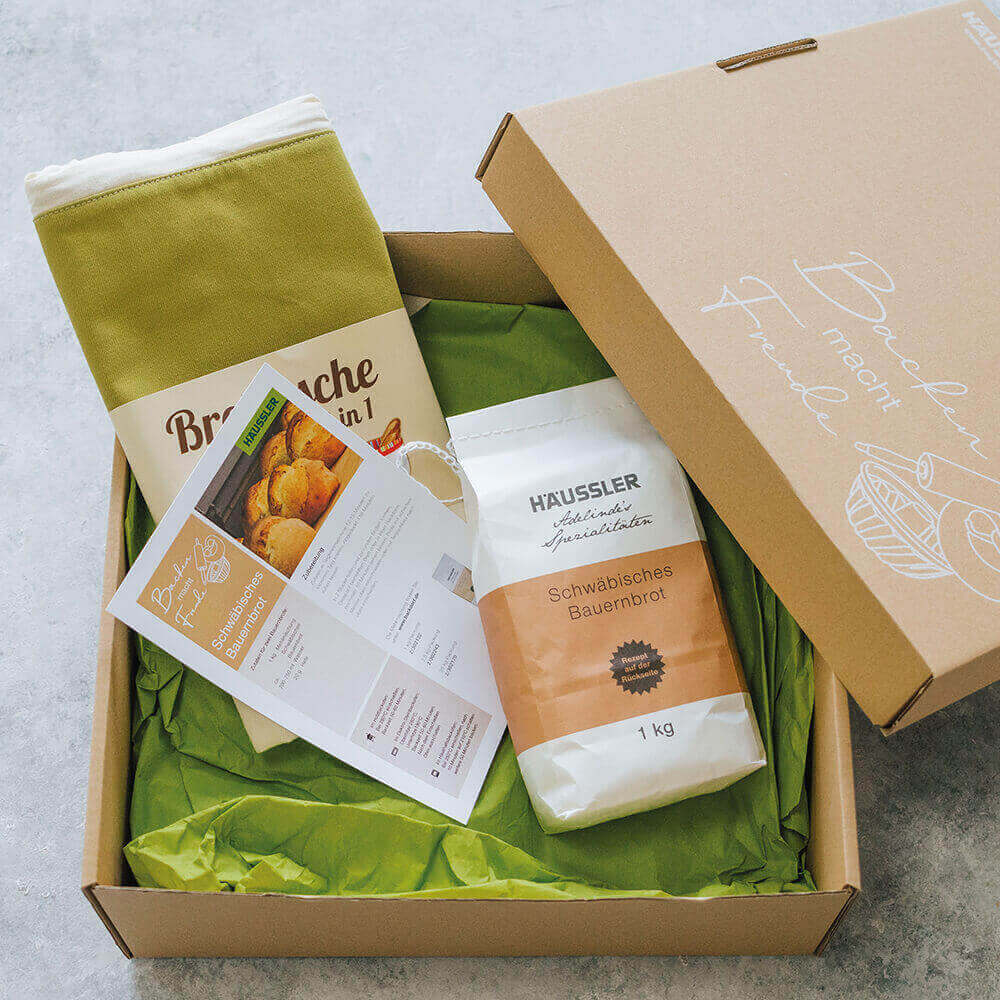 In einem braunen Karton befindet sich grünes Füllpapier sowie eine Backmischung Schwäbisches Bauernbrot 1 Kilogramm Eine Stoffbrottasche in grün und ein Rezept für ein Schwäbisches Bauernbrot