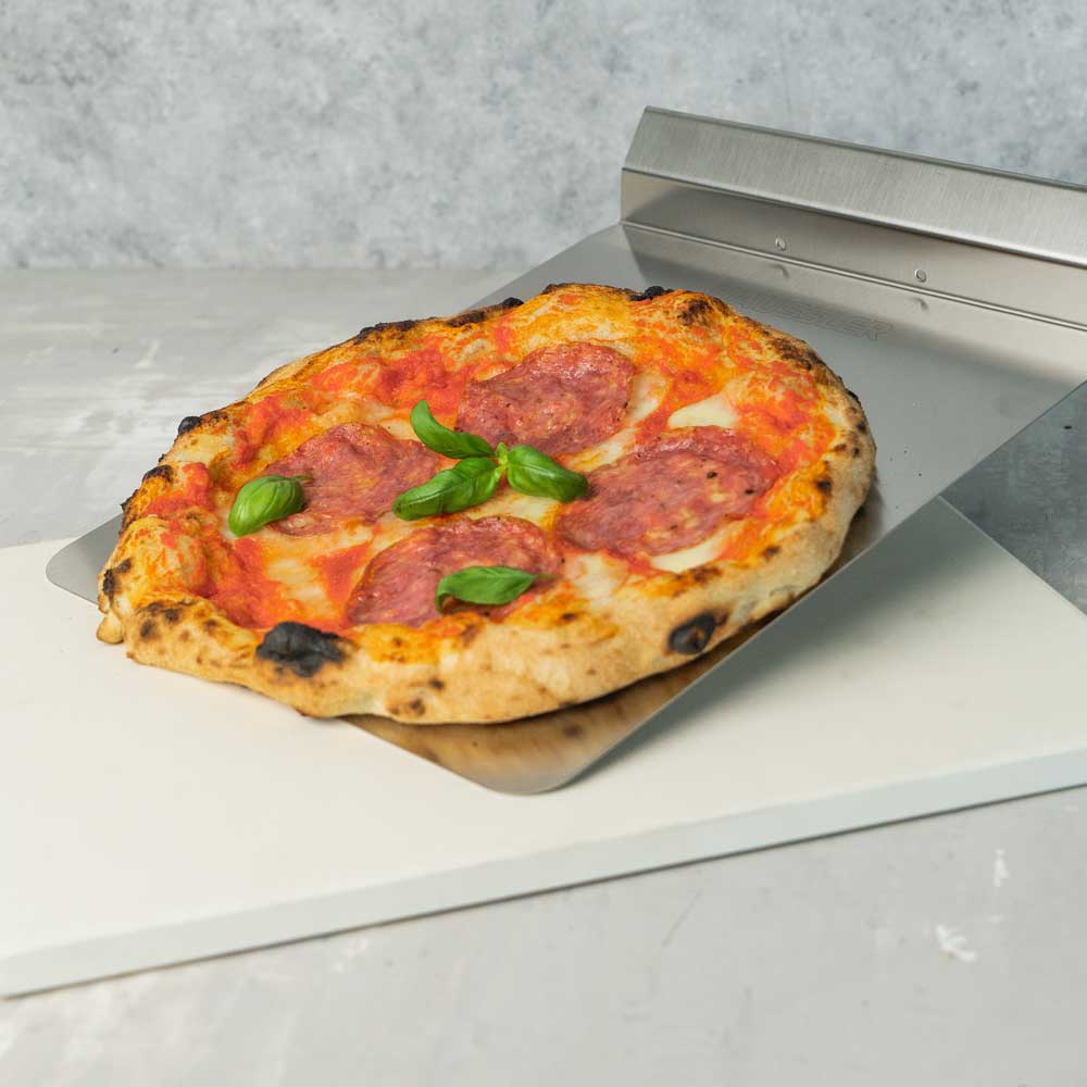weißer Pizzastein, darauf der Backschieber mit Handgriff und eine Salami-Pizza