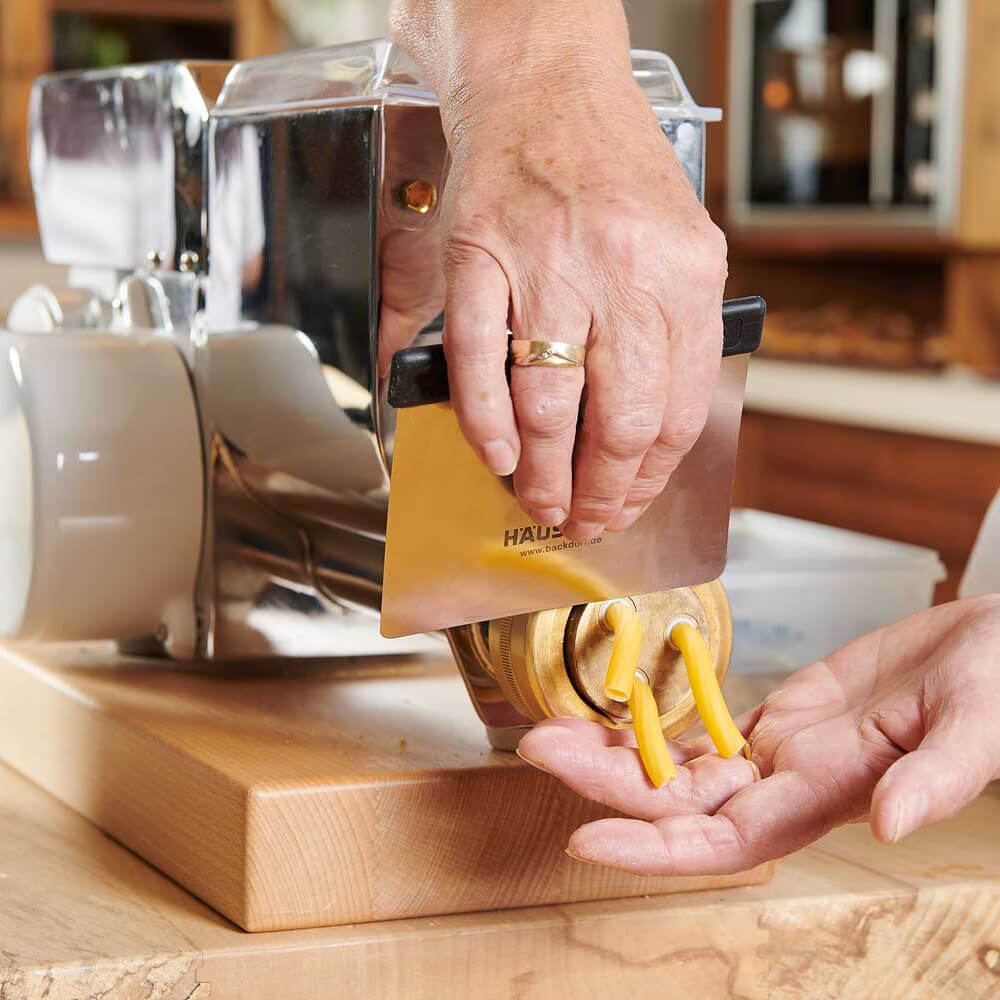 Hände die mit dem Teigabstecher Nudeln an einer Matrize der Nudelmaschine abstechen 