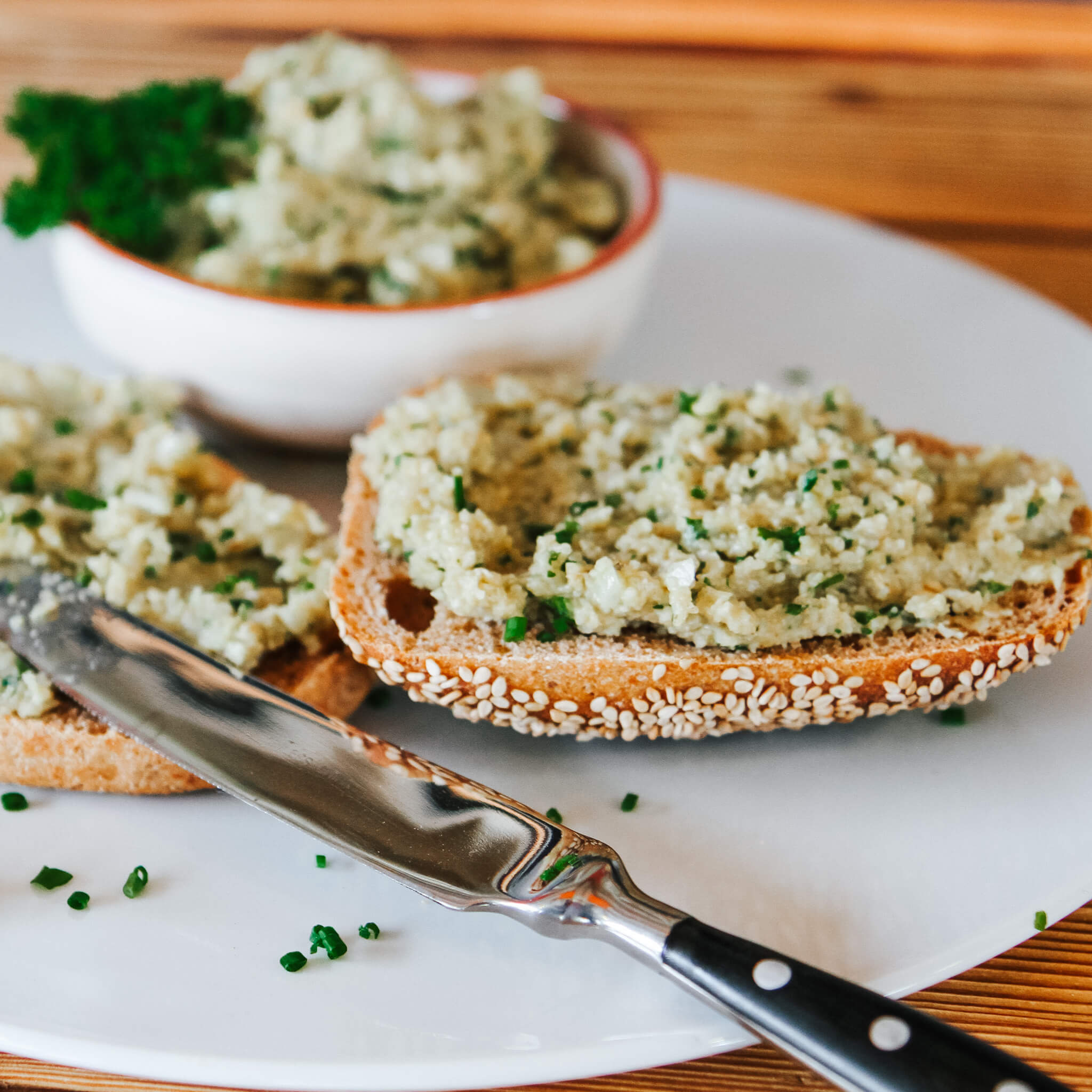 Rezept für Grünkerncreme Grünkerncreme mit Schnittlauch auf einem Körner Brötchen serviert auf einem weißen Teller mit einem Messer serviert