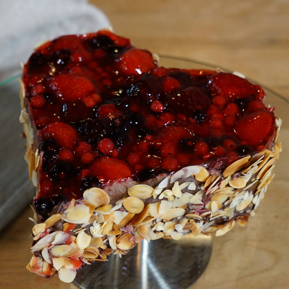 fruchtige Schokotorte in Herzform mit verschieden Beeren als Belag und Mandelblättchen verziert auf einer Glas Kuchenplatte serviert