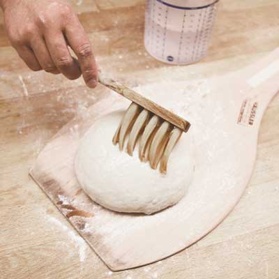 Ein ausgeformtes Brot liegt auf einem Backschieber und wird mit einem Brotpinsel mit wasser bestrichen. Das Brot ist noch nicht gebacken