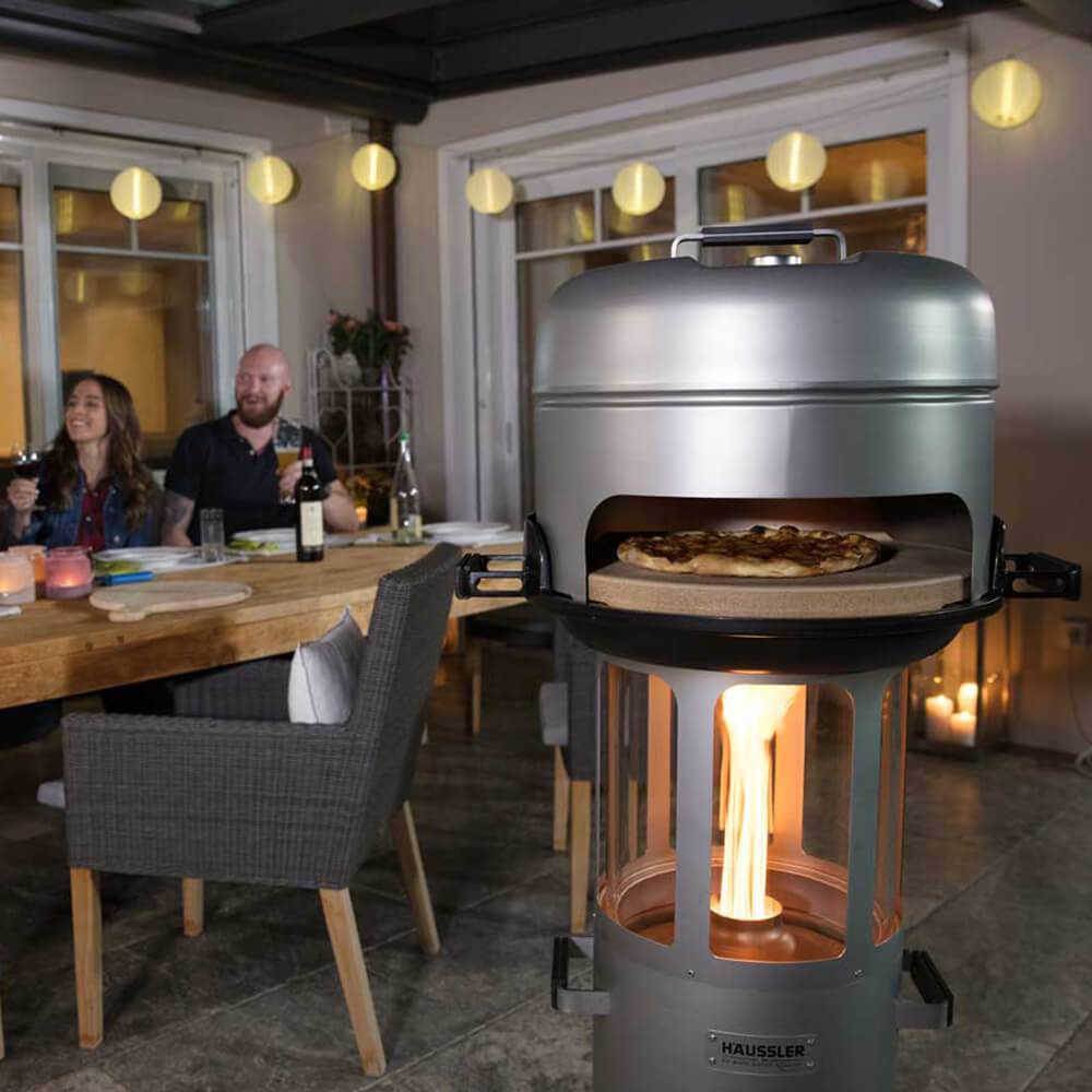 Pelettgrill mit einem Pizzastein, einem Zwischenring und einem Grilldeckel als Grillaufsatz. Im Hintergrund befindet sich eine Terrasse, die mit einer Lichterkette dekoriert ist.