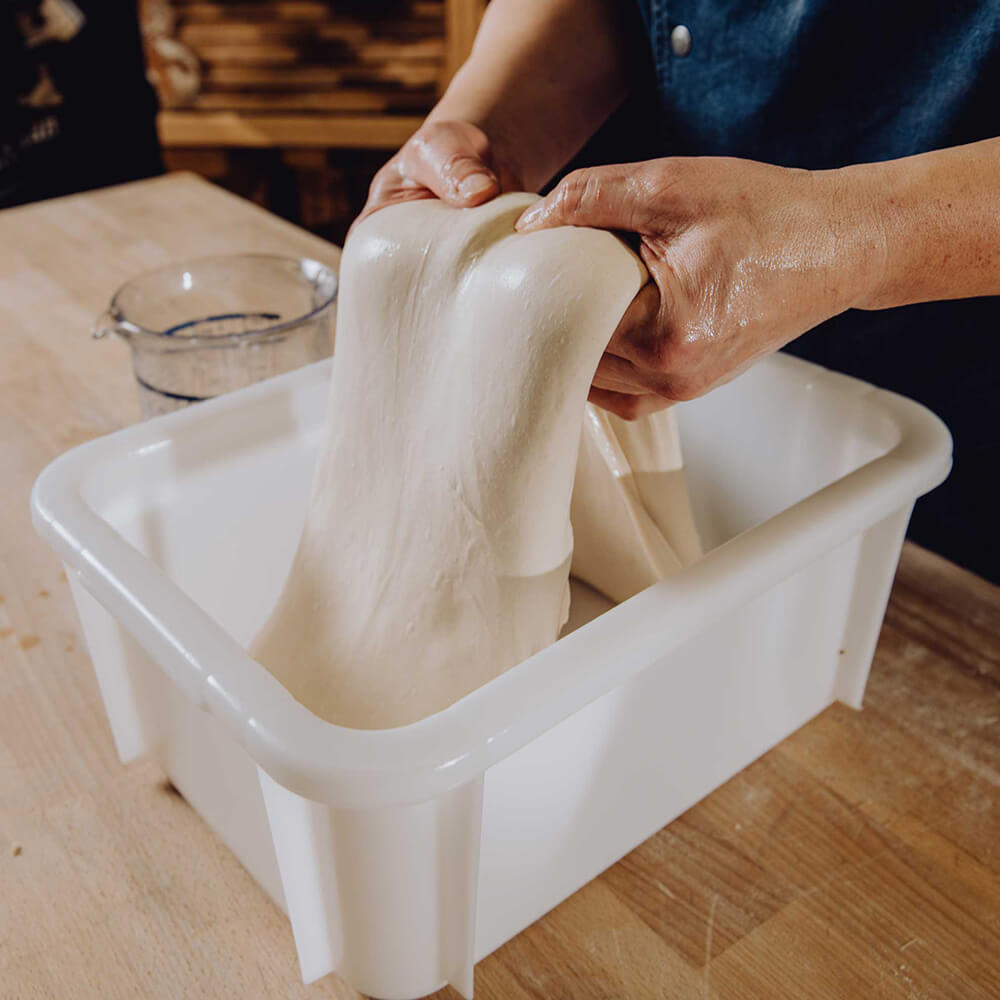 Ein Teig wird in einer Teigwanne gefalltet. Die Teigwanne steht auf einer braunen Arbeitsplatte