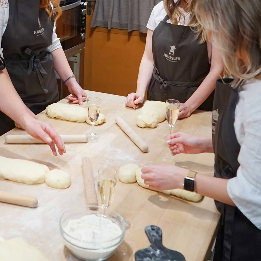 Häussler Schürze Arbeitstisch mit Teiglingen und Back-Werkzeug. 3 Personen die eine Häussler-Schürze tragen und am Arbeitstisch stehen.