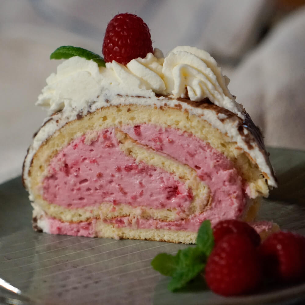 Rezept für Fruchtige Biskuitroulade 1 Stück Biskuitroulade gefüllt mit einer pinken Himbeersahnefüllung, dekoriert mit Sahne und einer Himbeere auf einem grauen Teller