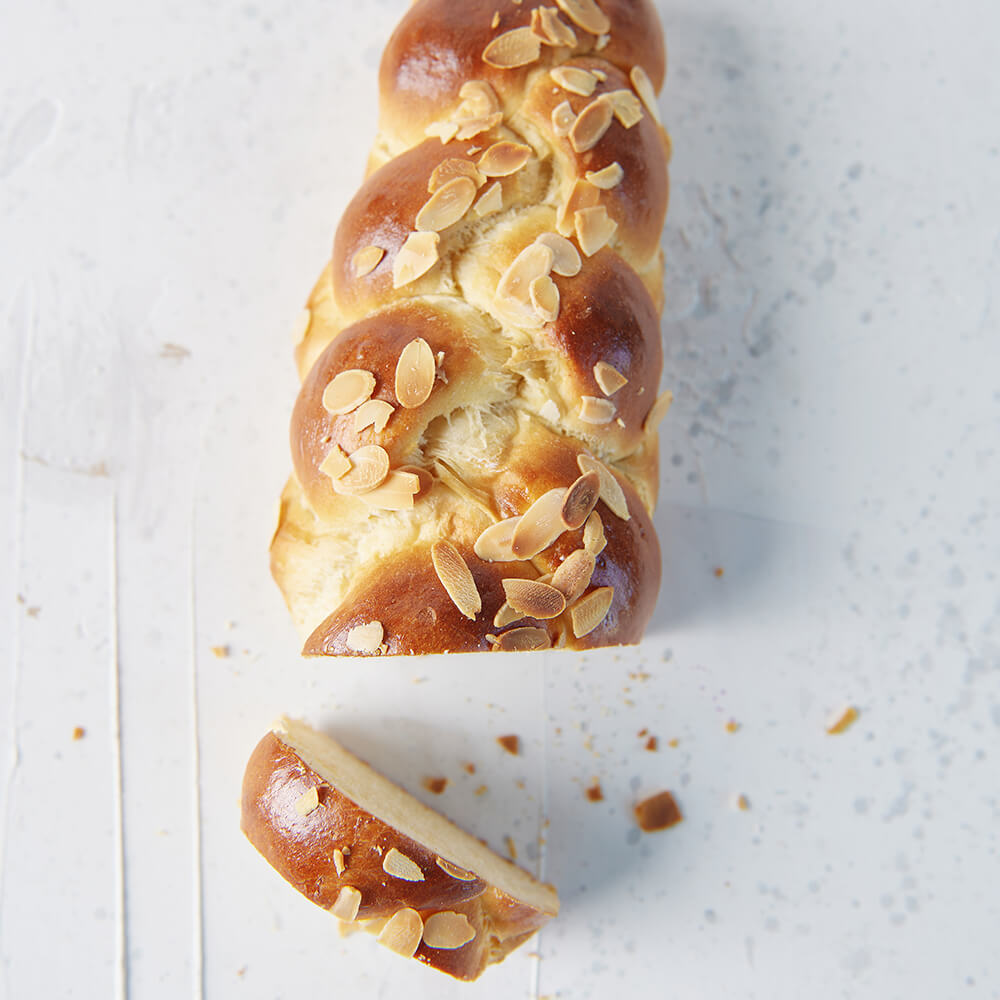 Rezept für Adelindes feiner Zopf leckere Hefezopf auf einem hellen Hintergrund, bestreut mit Mandelblättchen. Ein Stück wurde abgeschnitten