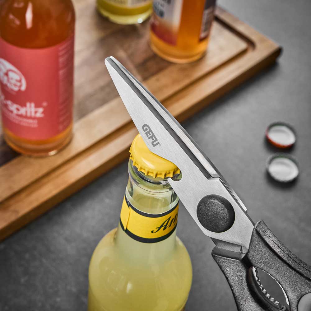 Allzweckschere Die Schere umgreift den Kronkorken einer Flasche, um diese zu öffnen. Im Hintergrund stehen noch weitere Flaschen und liegen weitere Kronkorken auf einer Arbeitsplatte.
