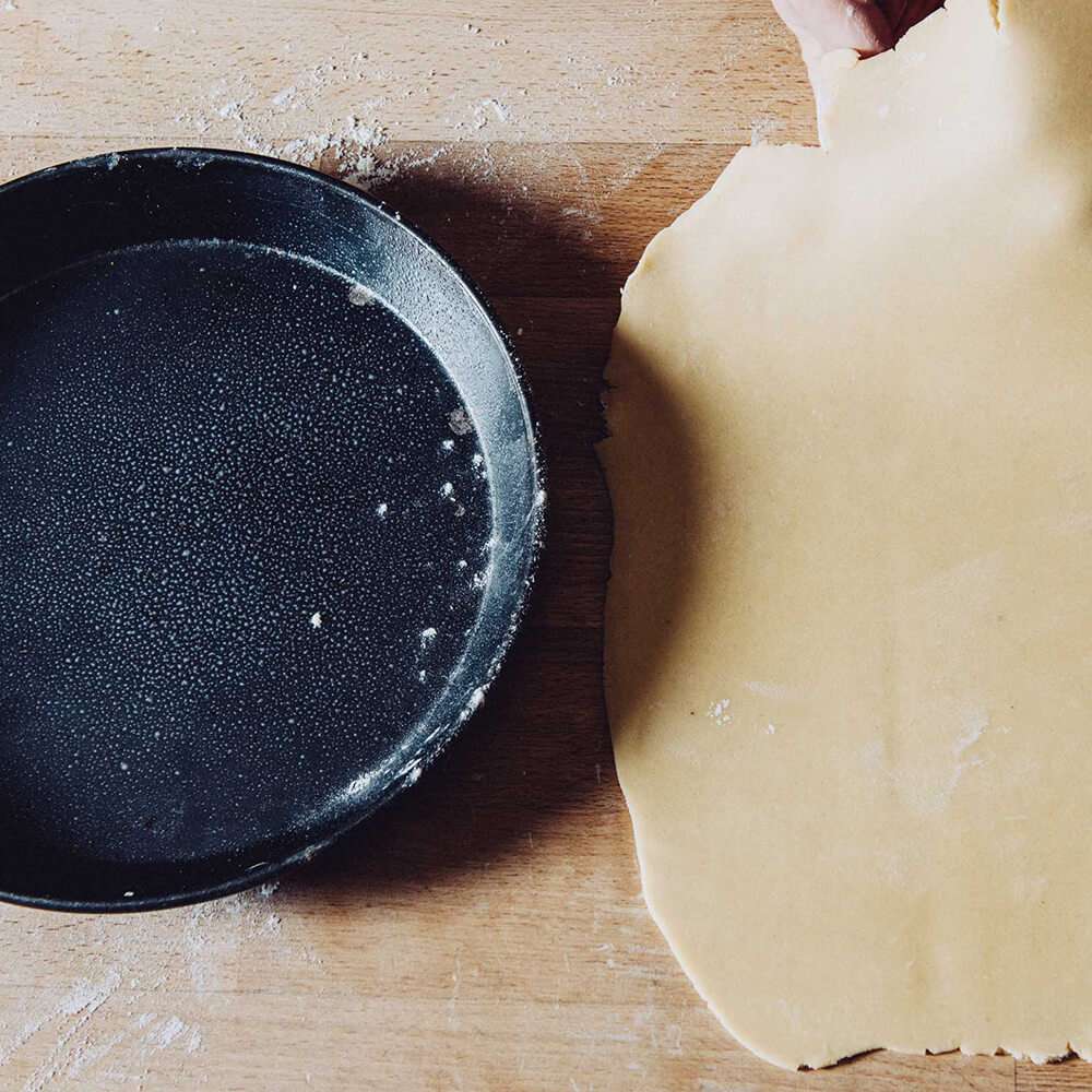 Links im Bild ist eine Blauglanzrundform abgebildet. auf der rechten Seite liegt ein ausgerollter Teig auf einer braunen Arbeitsfläche