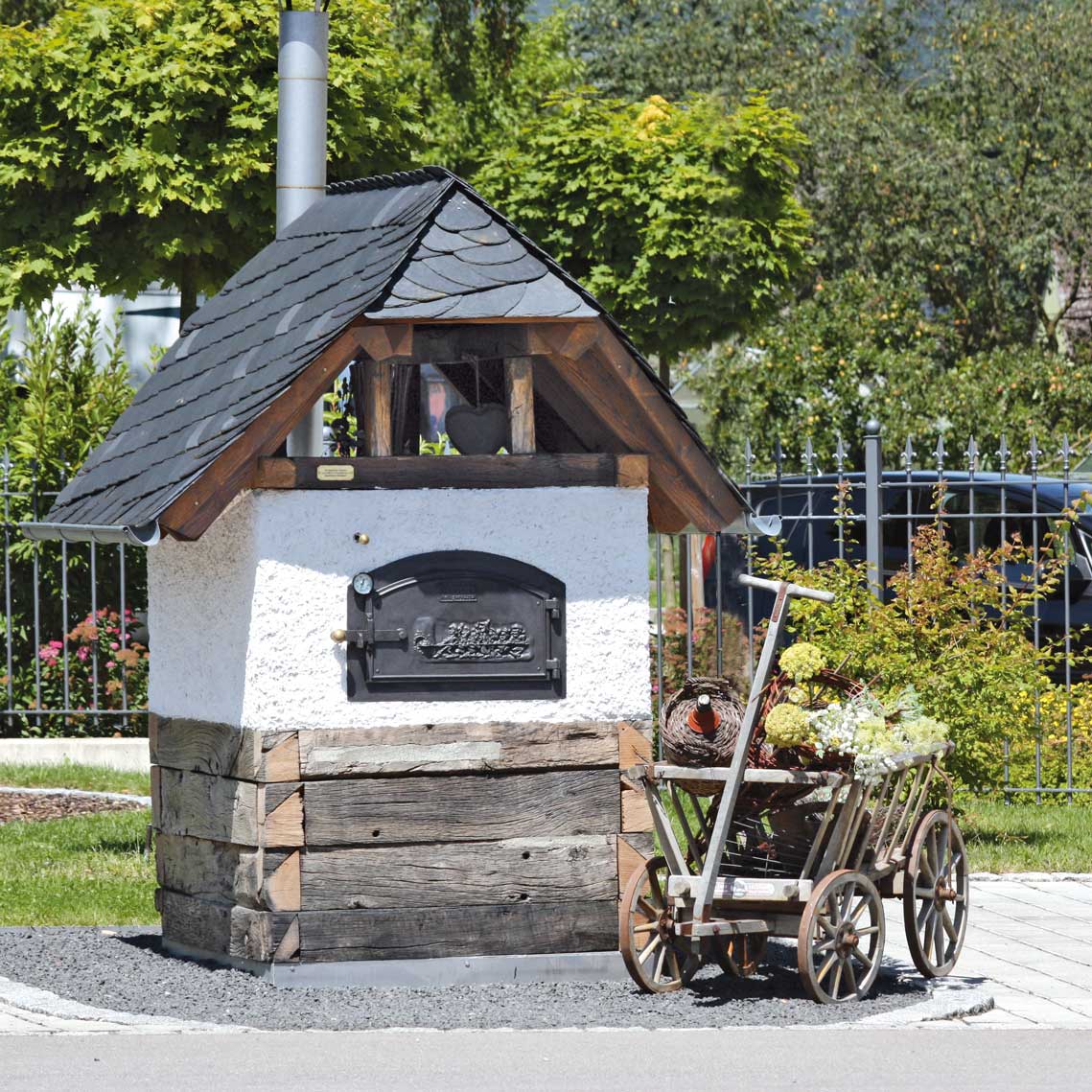 Aufgebauter HABO Bausatz zu einem schönen Backhäuschen mit einigen Bäumen im Hintergrund und einem Leiterwagen schräg vor dem Ofen.