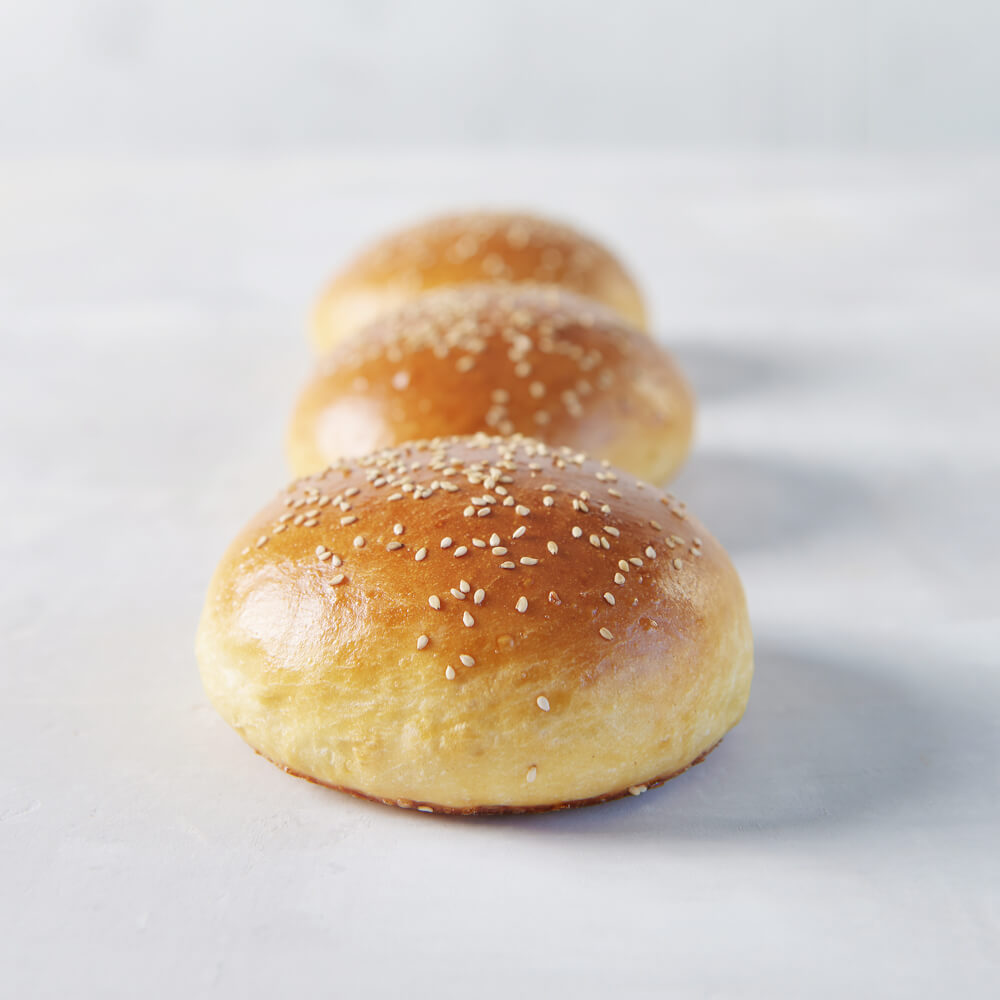 Drei fluffige Burger Buns mit Sesam bestreut vor einem hellgrauen Hintergrund