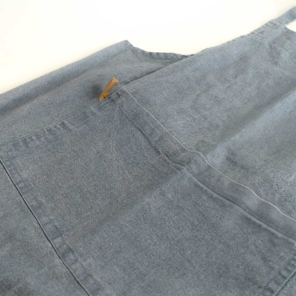 Schürze "MYRNA" blau Auf dem Bild befindet sich ein Ausschnitt einer blauen Schürze auf dem zu erkennen ist, das die schürze eine Tasche besitzt.