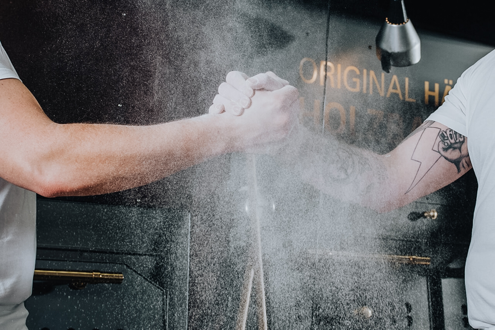 Zwei mit Mehl bestäubte Männerfäuste schlagen kräftig gegeneinander, dabei wirbelt feiner Mehlstaub durch die Luft - Symbol für Handwerk und Teamgeist.