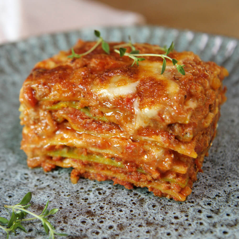 Rezept für Lasagne mit Bolognese ein Stück geschichtete Lasagne mit Bolognese und Bechamelsoße auf einem grauen Teller mit ein paar frischen Kräutern serviert
