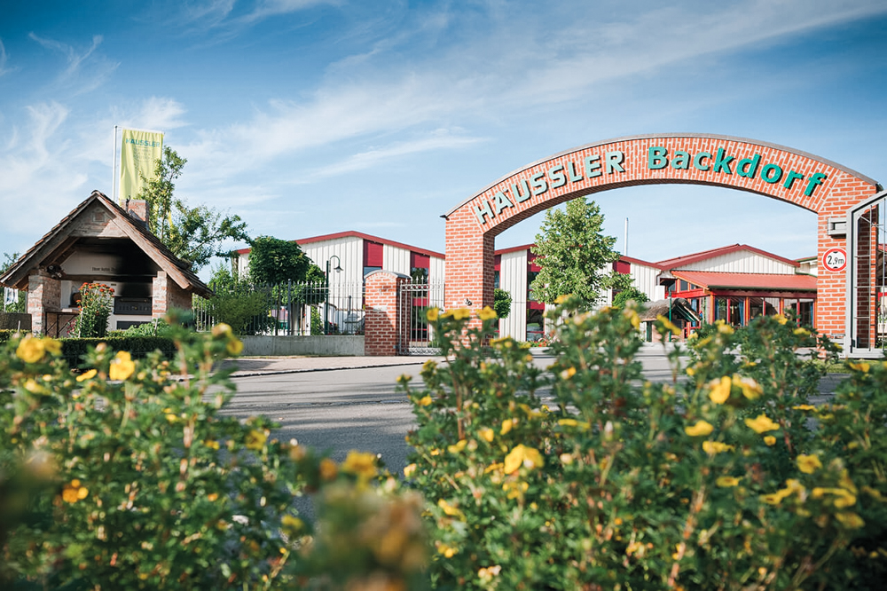 Aussenansicht des Häussler Backdorfs mit traditioneller Architektur, großen Eingangstor, Holz- und Steinelementen, umgeben von grüner Landschaft und einladender Atmosphäre.