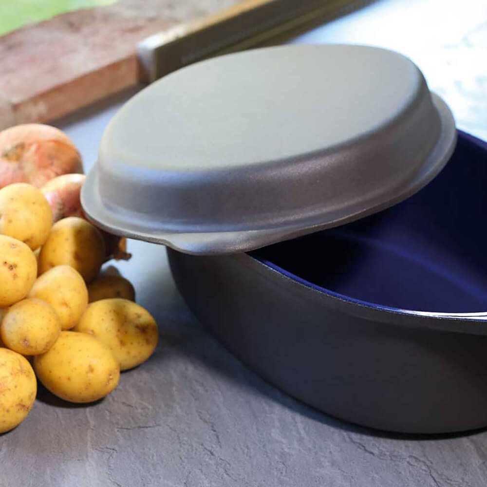 Auf dem Bild befindet sich ein Gussbräter mit zwei Griffen und einem Deckel. Daneben liegen Kartoffeln auf einer grauen Steinplatte.