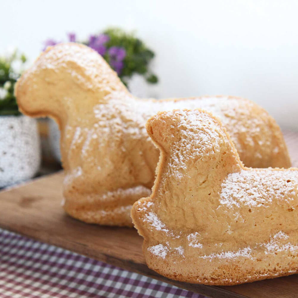 2 Osterlämmer nebeneinandersitzend auf einem Holzbrett. Im Hintergrund befinden sich zwei Blumensträuße. Eines der Osterlämmer hat den Kopf seitlich gerichtet und das andere Osterlamm schaut gerade aus
