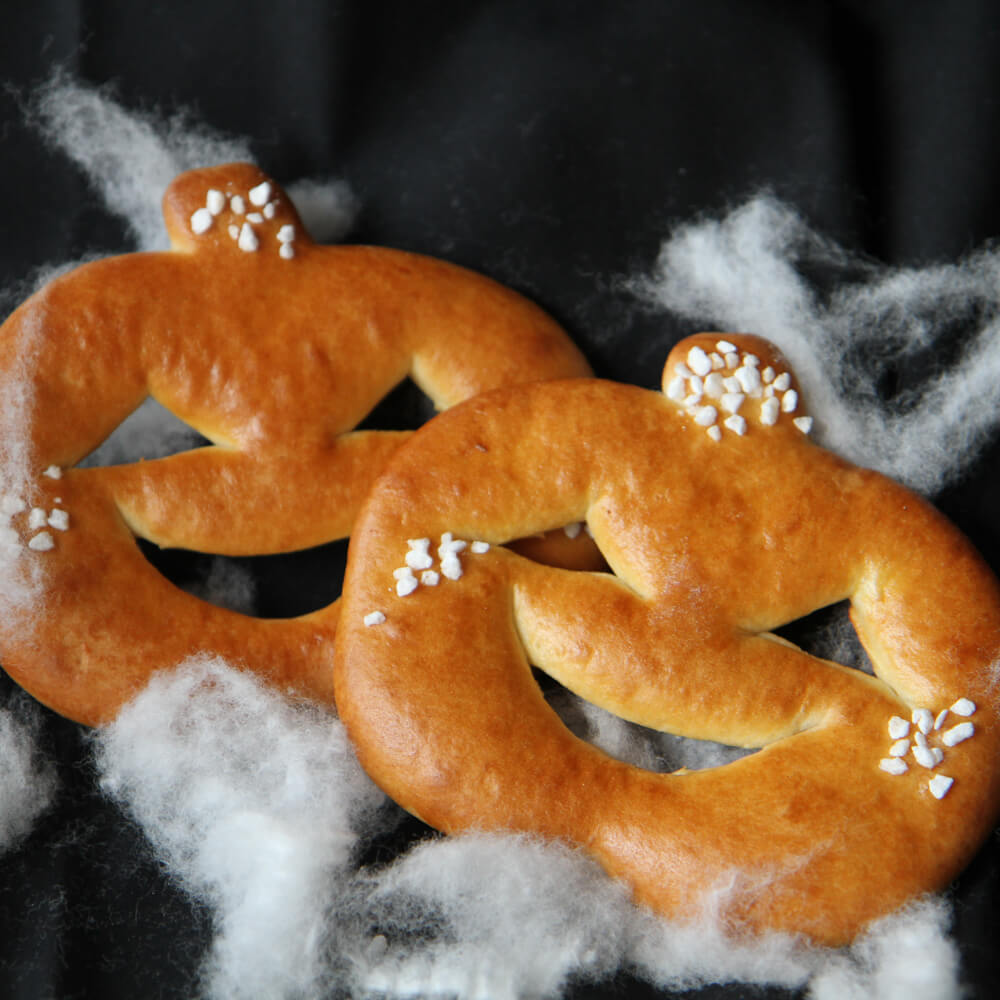 Rezept für Kürbis-Ausstecher 2 Kürbis-Austecher aus Hefezopfteig verziert mit Hagelzucker auf einem schwarzen Tuch mit weißer Watte