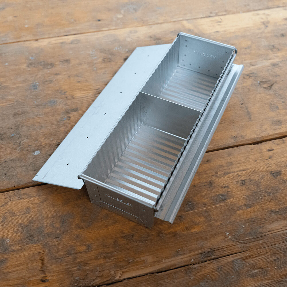 Mittlere Toastbrotform aus aluminiertem Stahl mit geöffnetem Deckel auf einem Holz-Untergrund. Die Abtrennung in der Form ist ersichtlich.